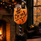 A Halloween-themed wine glass decoration displayed on a wooden table in a cozy living room setting with a fireplace. The stem is black and white striped, decorated with a large black bow at the base, and the bowl contains orange liquid with the text 'DON'T Drink and FLY!' along with small witch and ghost figurines.